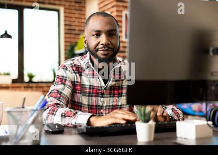 Focused african american entrepreneur works from home on desktop computer, reviewing marketing data and updating company financial reports before deadline. Serious, goal driven remote worker. Stock Photo