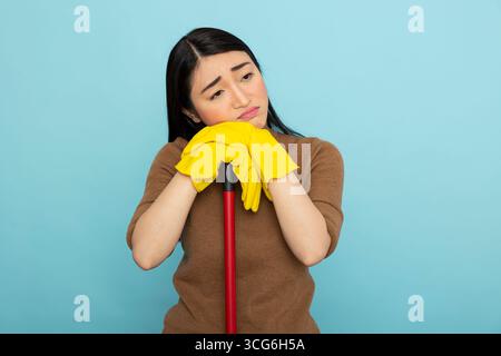 Bored female janitor leaning on mop, overwhelmed by cleaning duties. Exhausted asian housekeeper resting on broomstick, drained from repetitive household chores and long hours of service. Stock Photo