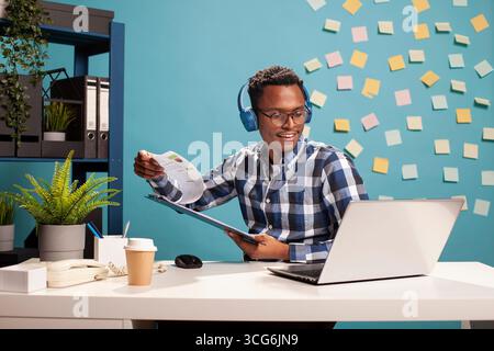 Businessman using wireless headphones and researching marketing strategies at his desk. Smiling office worker with clipboard in hand, enjoying some music as he reviews project ideas on his laptop. Stock Photo
