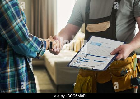 Caucasian middle aged man wearing tool belt holding invoice clipboard shaking hands with unseen young adult Caucasian woman in home interior, confirming service completion Stock Photo