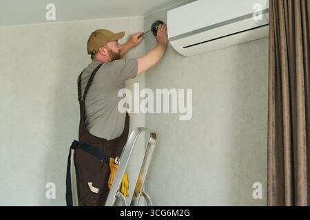 Caucasian middle aged man standing on ladder installing security camera near air conditioning unit, focusing on precise placement and adjustment of surveillance equipment Stock Photo