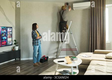 Caucasian middle aged man standing on ladder installing air conditioning unit while Caucasian young adult woman observing in modern living room Stock Photo