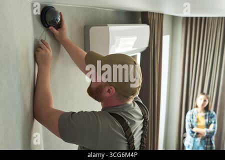 Caucasian middle aged man installing security camera on wall while young Caucasian woman standing in background observing process in modern residential interior Stock Photo