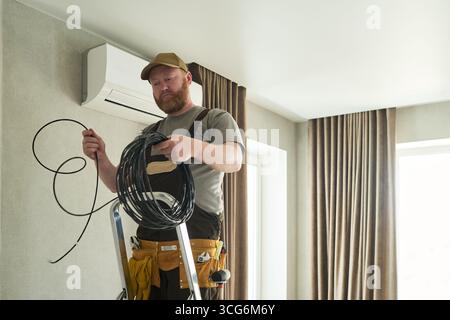 Caucasian middle aged man standing on ladder installing air conditioning unit, holding coiled cable and preparing for electrical work in modern residential interior Stock Photo