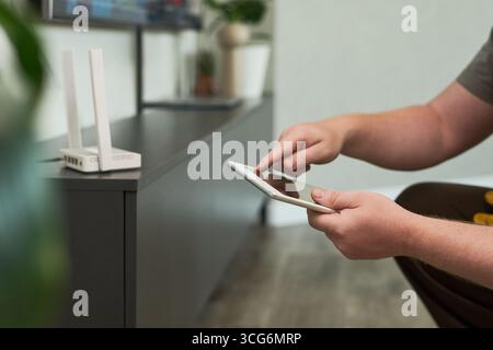 Caucasian middle aged man configuring wireless router using digital tablet, sitting near modern cabinet, focusing on device setup process, hands interacting with touchscreen Stock Photo