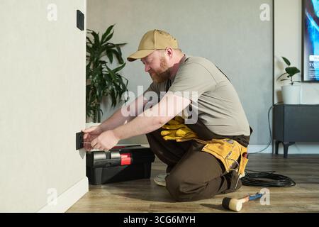 Caucasian middle aged man kneeling on floor installing electrical outlet using screwdriver, tool belt around waist, toolbox and hammer nearby, working in modern residential interior Stock Photo