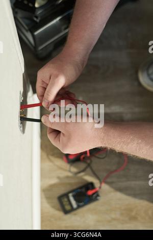 Caucasian middle aged man using digital multimeter to test electrical wiring in wall socket, hands holding probes while measuring voltage, device resting on wooden floor Stock Photo