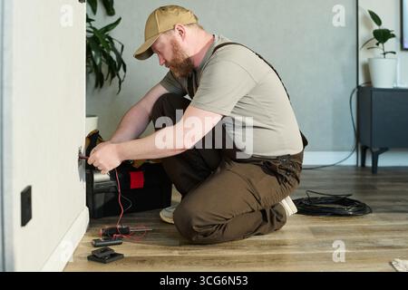 Middle aged Caucasian man kneeling on floor repairing electrical outlet using tools and wires, focusing on task with toolbox and coiled cable nearby in modern indoor setting Stock Photo