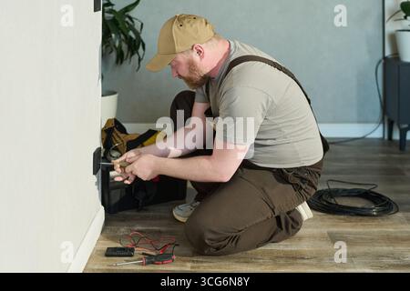 Caucasian middle aged man kneeling on floor repairing electrical outlet using tools, wearing work uniform and cap, tool bag and coiled cable placed nearby, working indoors Stock Photo