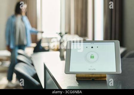 Tablet displaying security system interface with lock icon on kitchen counter, Caucasian young adult woman standing in background adjusting object, home interior visible Stock Photo