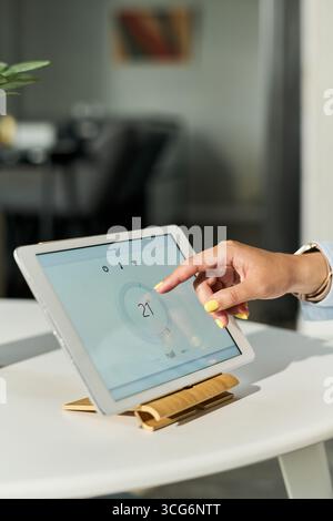 Caucasian young adult woman adjusting smart thermostat on tablet device with finger, closeup of hand interacting with digital temperature control inte Stock Photo