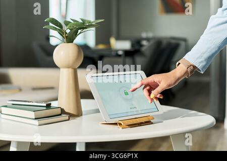Caucasian young adult woman using tablet device on table, unlocking screen with finger, modern home interior in background, partial view showing hand Stock Photo