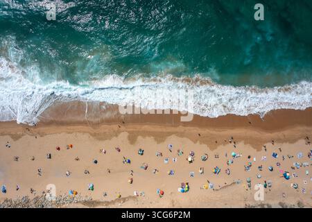 Overhead drone shot of anonymous tourist and colorful parasols on sandy seashore, showing people enjoying summer vacations at beach during sunny day  Stock Photo