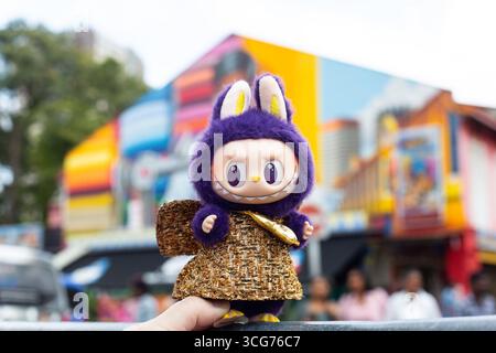 Purple colour Labubu in gold shiny dress posing against wall mural, street at Little India, Singapore. Stock Photo