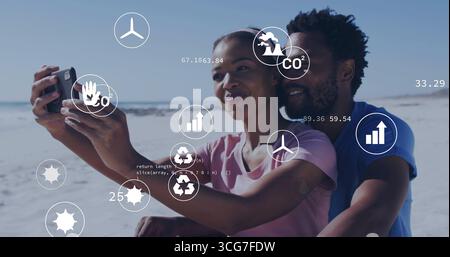 Couple taking selfie and sitting closely on sandy shoreline, with phone and floating eco data icons Stock Photo