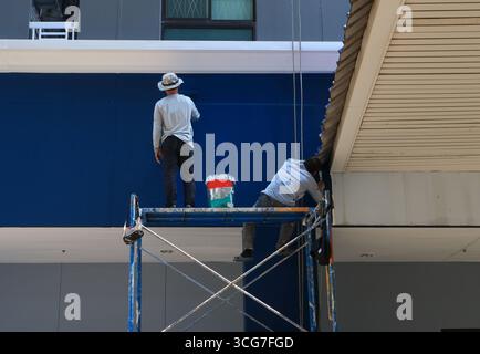 Bangkok, Thailand - 5 April, 2023 : Two men workers are painting tall building wall with paint roller, one of them standing on scaffolding while the o Stock Photo