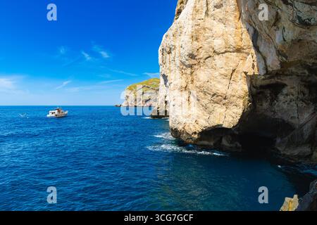 Steep limestone cliffs at Capo Caccia, Sardinia, with a white boat floating on deep blue sea and a bright summer sky overhead. Stock Photo