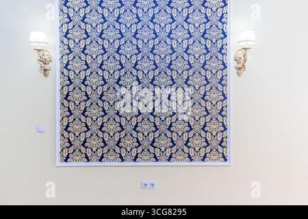 A wall with a blue and gold patterned wallpaper. The wallpaper is framed and has a gold trim. There are two light fixtures on the wall, one on the lef Stock Photo