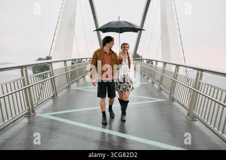 Joyful couple strolls beneath a black umbrella, enjoying the rain together in a scenic park. Stock Photo