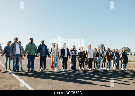 A diverse group of adults stands side by side, joining hands in unity outdoors. They are positioned on a wide open road, with urban buildings visible Stock Photo