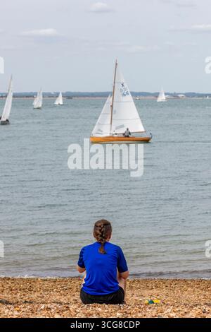 young woman sitting alone on a shingle beach watching the cowes week yacht racing on the isle of wight uk. Young woman watching boats from beach. Stock Photo