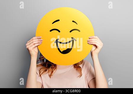 Happy emoji. Girl holds a yellow emoticon with a smile isolated on a gray background. Happy emoji Stock Photo