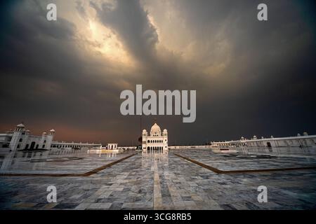 View of a serene white temple complex reflecting in the wet marble ground under dramatic, brooding skies, Kartarpur, Punjab, Pakistan. Stock Photo