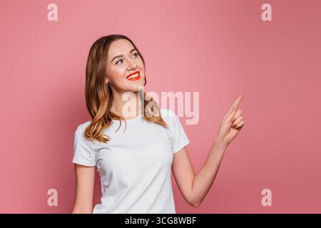 Young caucasian brown-haired woman with red lipstick smiling and pointing finger at copy space isolated on pink background Stock Photo