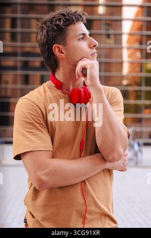 Young man in brown t-shirt with red headphones thinking outdoors in the city Stock Photo