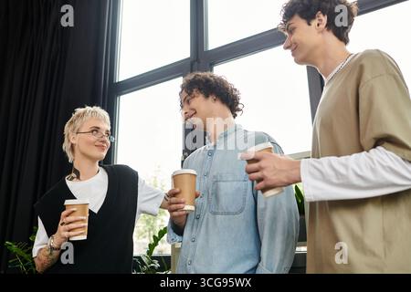 Three young professionals share stories and laughs over coffee in a vibrant office environment. Stock Photo