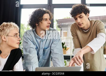 Three young professionals focus intently on a laptop, brainstorming ideas amid a bright office. Stock Photo