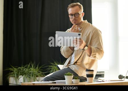 A young businessman wearing glasses thoughtfully engages with a digital tablet in his office. Stock Photo