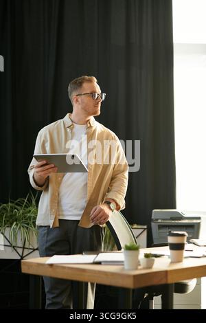 A focused young professional in a sunlit office holds a tablet, pondering work tasks. Stock Photo