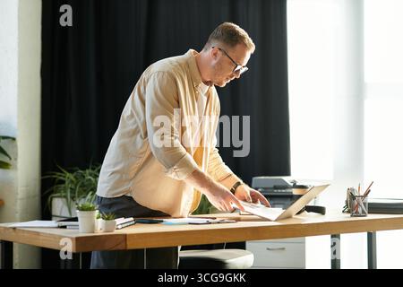 A young businessman diligently reviews documents while standing at his office desk. Stock Photo