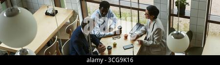 Header of three middle aged business professionals, including Black man and Caucasian man and woman, sitting at table discussing work, holding coffee cups, smartphone and documents on table Stock Photo