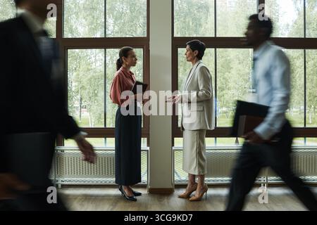 Two middle aged Caucasian women standing and talking in office hallway holding folders while two young adult men, one Black and one Caucasian, walking past in foreground Stock Photo