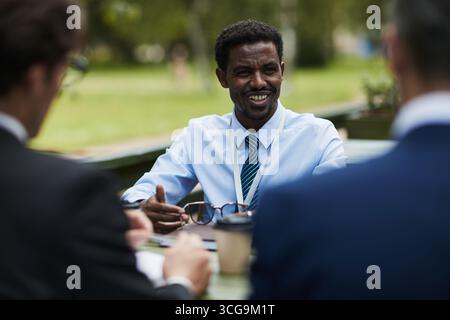 Black young man smiling while discussing business with two Caucasian male colleagues outdoors, holding eyeglasses and gesturing with hands, coffee cup and documents on table Stock Photo
