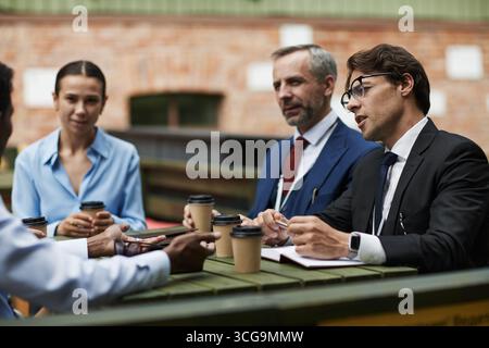 Diverse group of middle aged multiethnic business professionals discussing project outdoors, sitting at table with coffee cups, man in suit gesturing and speaking, woman and man listening attentively Stock Photo