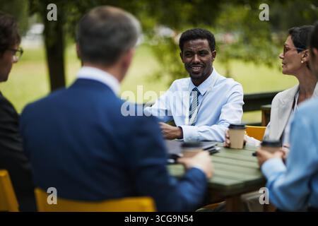 Black young man leading outdoor business meeting with multiethnic colleagues sitting around table discussing project, holding coffee cups, engaging in conversation Stock Photo