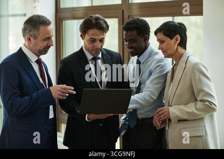 Group of middle aged multiethnic business professionals collaborating around laptop, discussing project details in modern office setting, focused on screen and engaged in teamwork Stock Photo