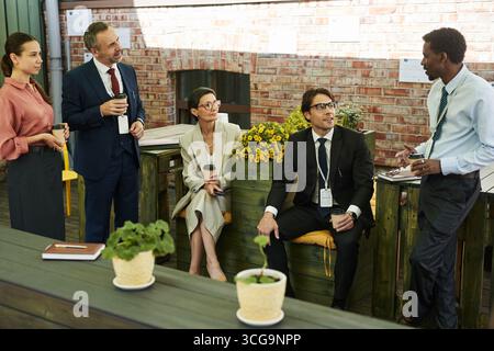 Group of middle aged and young multiethnic business professionals standing and sitting outdoors holding coffee cups and engaging in conversation during coffee break Stock Photo
