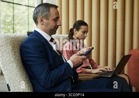 Middle aged Caucasian man using smartphone while sitting next to young woman working on laptop, both focused on digital devices in modern corporate office setting Stock Photo
