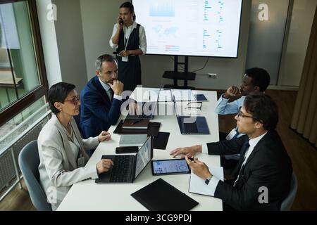 Diverse group of middle aged and young men and women working at table with laptops and documents, businesswoman standing in background talking on phone, large screen displaying charts Stock Photo
