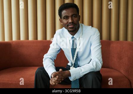 Portrait of Black man sitting on sofa, wearing business attire, looking into camera with confident expression, hands clasped, appearing as young professional in office setting Stock Photo