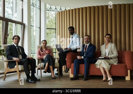 Group of middle aged and young business professionals including Caucasian man, woman, Black man, Asian woman sitting together in modern office setting, working and posing Stock Photo