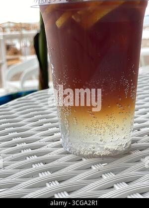 Iced tea in a clear plastic cup with lemon slices and bubbles, on woven table, seaside background. Mobile photo Stock Photo