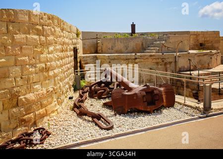 A large rusty sea anchor with a massive metal chain lies on the gravel in a historic fortress with steps and stone walls. Stock Photo