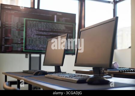 Empty computer lab featuring two desktop monitors and keyboards on desks with large screen displaying programming code in background, modern workspace setting for software development Stock Photo