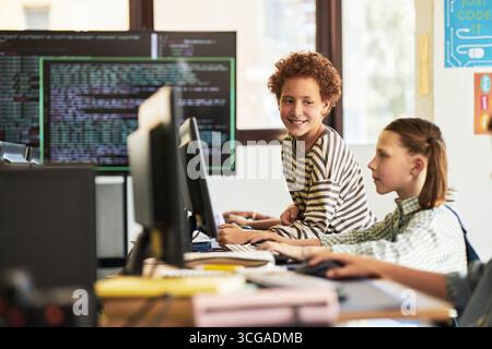 Two Caucasian teenagers sitting at desks using desktop computers in classroom, one teenager smiling and looking at classmate while programming code displayed on monitors in background Stock Photo
