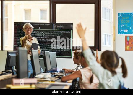 Senior Caucasian woman teaching group of diverse children in computer classroom, holding digital tablet while smiling and pointing at students raising hands in front of monitors Stock Photo